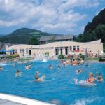 Öffentliches Freibad mit vielen Menschen, die im Wasser schwimmen und spielen, umgeben von Bäumen, Hügeln und einem Gebäude im Hintergrund.