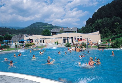 Öffentliches Freibad mit vielen Menschen, die im Wasser schwimmen und spielen, umgeben von Bäumen, Hügeln und einem Gebäude im Hintergrund.