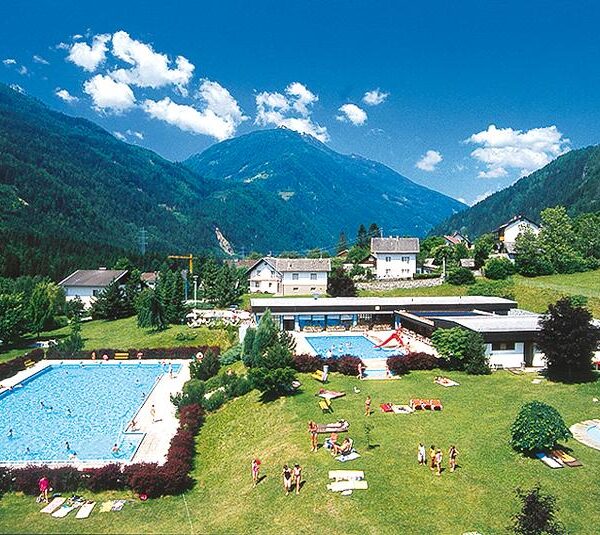 Freibadkomplex mit Menschen, die sich sonnen und schwimmen, umgeben von Gras, Gebäuden und grünen Bergen unter einem blauen Himmel.
