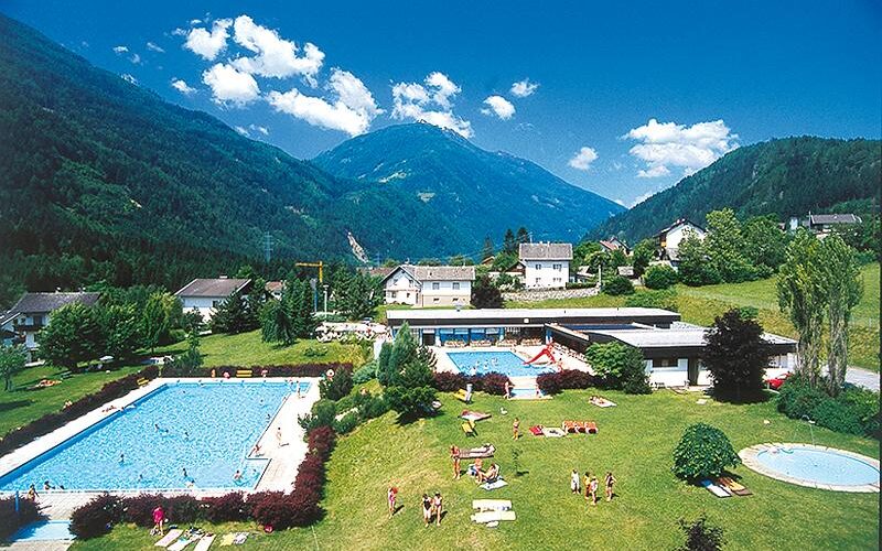 Freibadkomplex mit Menschen, die sich sonnen und schwimmen, umgeben von Gras, Gebäuden und grünen Bergen unter einem blauen Himmel.