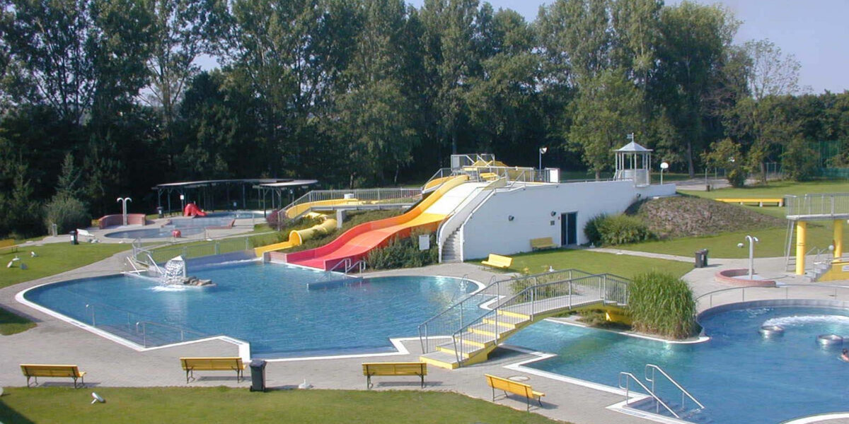 Freibadkomplex mit mehreren Becken, Wasserrutschen, Springbrunnen, Rasenflächen und Bänken, umgeben von Bäumen unter klarem Himmel.