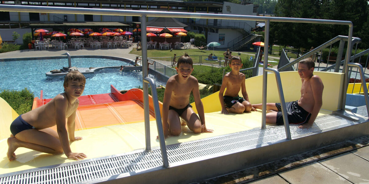 An einem sonnigen Tag sitzen vier Jungen in Badeanzügen oben auf einer Wasserrutsche in einem Freibad.