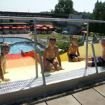 An einem sonnigen Tag sitzen vier Jungen in Badeanzügen oben auf einer Wasserrutsche in einem Freibad.