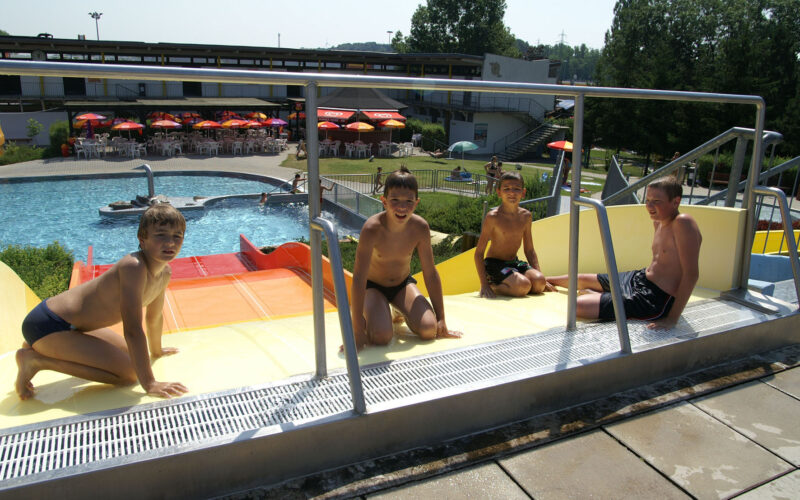 An einem sonnigen Tag sitzen vier Jungen in Badeanzügen oben auf einer Wasserrutsche in einem Freibad.