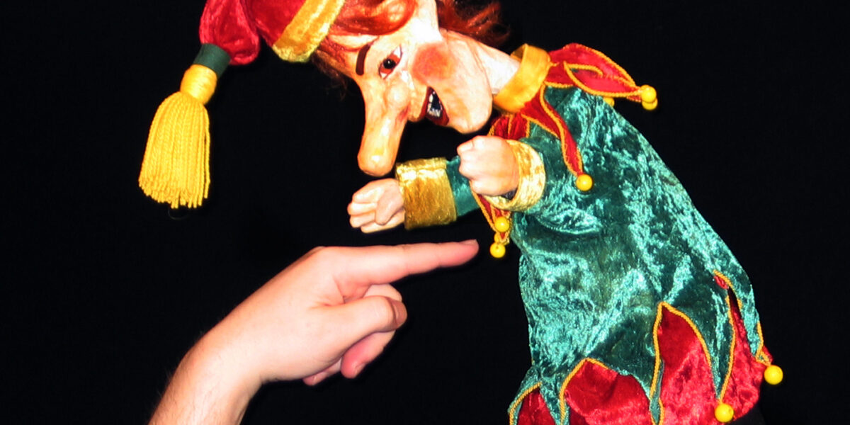 Eine Puppe in einem grün-roten Narrenkostüm wird vor einem schwarzen Hintergrund mit dem Finger einer Person berührt.