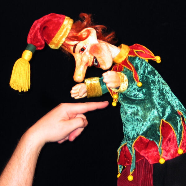 Eine Puppe in einem grün-roten Narrenkostüm wird vor einem schwarzen Hintergrund mit dem Finger einer Person berührt.