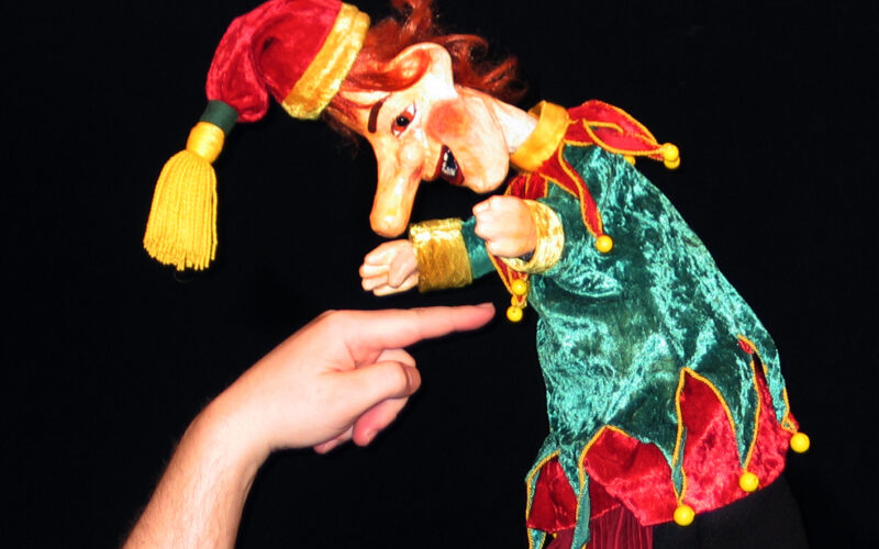 Eine Puppe in einem grün-roten Narrenkostüm wird vor einem schwarzen Hintergrund mit dem Finger einer Person berührt.