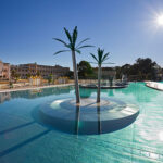 Außenschwimmbad mit zwei künstlichen Palmen auf einer kreisförmigen Plattform in der Mitte, umgeben von klarem Wasser und Gebäuden im Hintergrund unter strahlender Sonne.