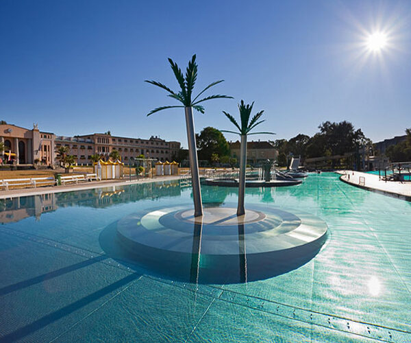 Außenschwimmbad mit zwei künstlichen Palmen auf einer kreisförmigen Plattform in der Mitte, umgeben von klarem Wasser und Gebäuden im Hintergrund unter strahlender Sonne.
