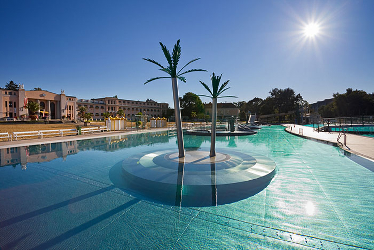 Außenschwimmbad mit zwei künstlichen Palmen auf einer kreisförmigen Plattform in der Mitte, umgeben von klarem Wasser und Gebäuden im Hintergrund unter strahlender Sonne.