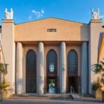 Symmetrisches Gebäude mit hohen Bogenfenstern, großen Säulen, Palmen, Fahnen auf jeder Seite und weißen Statuen auf dem Dach unter einem klaren blauen Himmel.