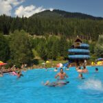 Freibad mit schwimmenden und sich entspannenden Menschen, einer blauen Spiralrutsche und bewaldeten Hügeln im Hintergrund unter einem teilweise bewölkten Himmel.