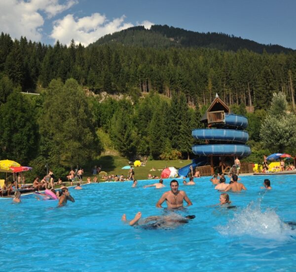 Freibad mit schwimmenden und sich entspannenden Menschen, einer blauen Spiralrutsche und bewaldeten Hügeln im Hintergrund unter einem teilweise bewölkten Himmel.