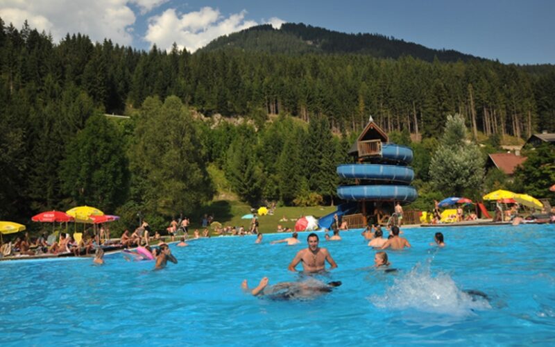 Freibad mit schwimmenden und sich entspannenden Menschen, einer blauen Spiralrutsche und bewaldeten Hügeln im Hintergrund unter einem teilweise bewölkten Himmel.