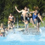 Eine Gruppe von Kindern in Badeanzügen springt in ein Schwimmbecken und verursacht große Spritzer, während andere an einem sonnigen Tag vom Beckenrand aus zusehen.