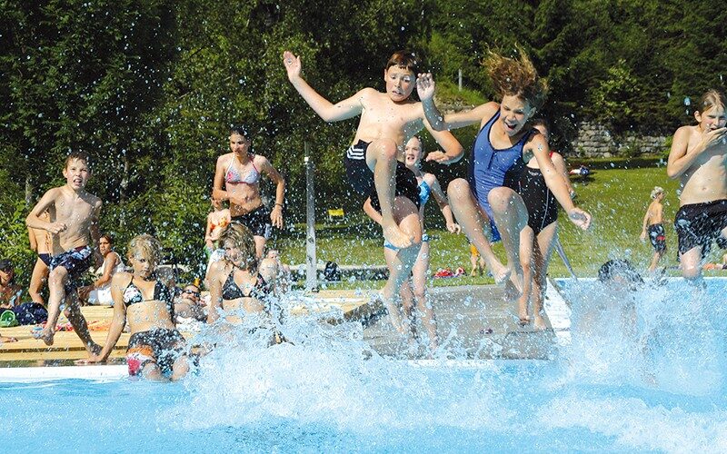 Eine Gruppe von Kindern in Badeanzügen springt in ein Schwimmbecken und verursacht große Spritzer, während andere an einem sonnigen Tag vom Beckenrand aus zusehen.