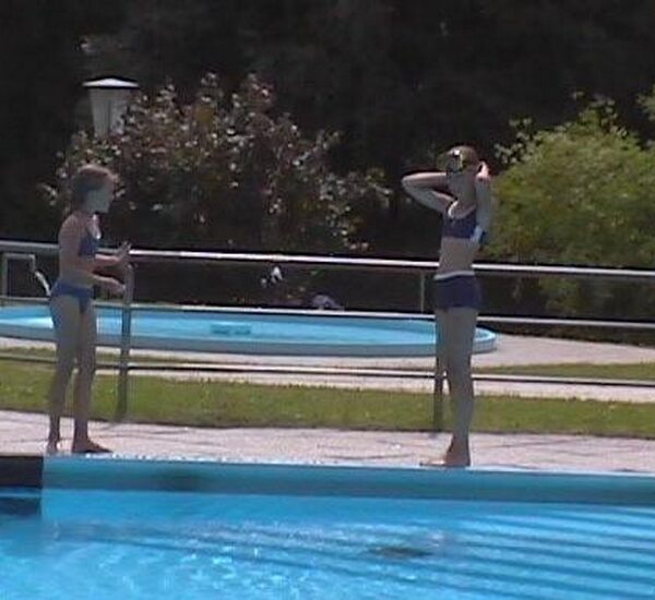 Zwei Kinder in Badeanzügen stehen am Rande eines Schwimmbeckens, wobei eines von ihnen seine Schwimmbrille zurechtrückt. Im Hintergrund sind ein rundes Planschbecken und Grünflächen zu sehen.