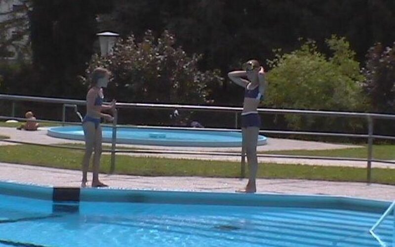 Zwei Kinder in Badeanzügen stehen am Rande eines Schwimmbeckens, wobei eines von ihnen seine Schwimmbrille zurechtrückt. Im Hintergrund sind ein rundes Planschbecken und Grünflächen zu sehen.