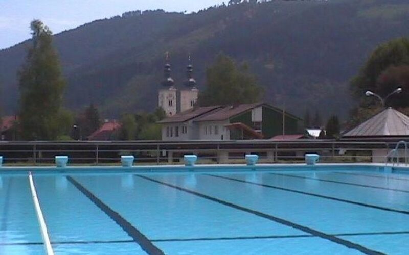 Freibad mit Bahnmarkierungen im Vordergrund, mit Gebäuden, Bäumen und Hügeln im Hintergrund unter einem bewölkten Himmel.