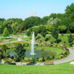 Ein Landschaftspark mit einem runden Brunnen, umliegenden Blumenbeeten, Bänken und Spazierwegen; im Hintergrund sind Bäume und ein Baukran zu sehen.