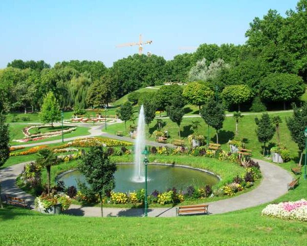 Ein Landschaftspark mit einem runden Brunnen, umliegenden Blumenbeeten, Bänken und Spazierwegen; im Hintergrund sind Bäume und ein Baukran zu sehen.