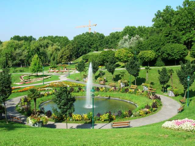 Ein Landschaftspark mit einem runden Brunnen, umliegenden Blumenbeeten, Bänken und Spazierwegen; im Hintergrund sind Bäume und ein Baukran zu sehen.