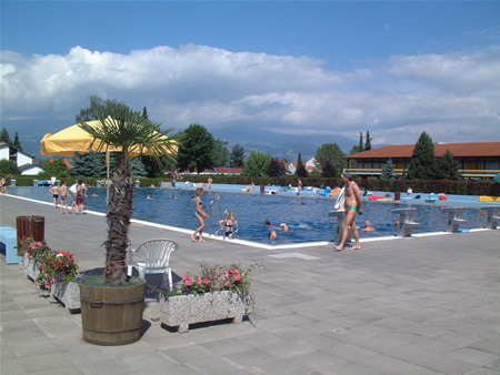 Außenschwimmbad mit schwimmenden und spazierenden Menschen, umgeben von Topfpflanzen und Stühlen, mit Gebäuden und Bergen im Hintergrund unter einem teilweise bewölkten Himmel.