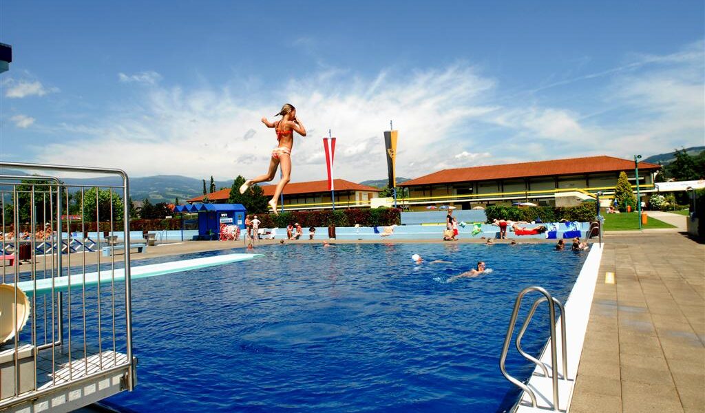 Eine Person springt in ein Freibad, während andere an einem sonnigen Tag um den Pool herum schwimmen und sich entspannen.