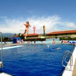 Eine Person springt in ein Freibad, während andere an einem sonnigen Tag um den Pool herum schwimmen und sich entspannen.