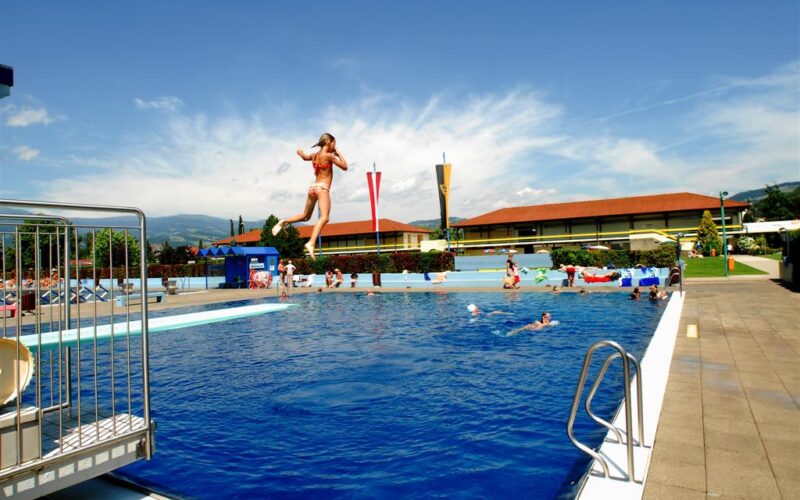 Eine Person springt in ein Freibad, während andere an einem sonnigen Tag um den Pool herum schwimmen und sich entspannen.