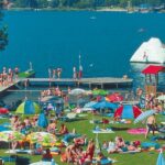 Belebter Strand am See mit Menschen, die sich im Gras sonnen, bunten Sonnenschirmen, einem Steg und Schwimmern; ein aufblasbarer Eisberg schwimmt im Wasser.