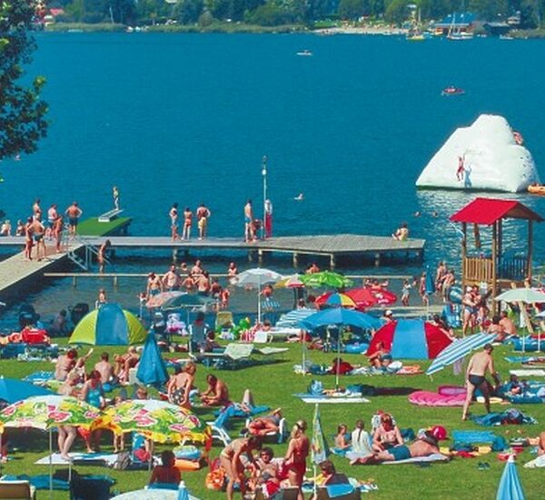 Belebter Strand am See mit Menschen, die sich im Gras sonnen, bunten Sonnenschirmen, einem Steg und Schwimmern; ein aufblasbarer Eisberg schwimmt im Wasser.