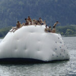 Eine Gruppe von Menschen sitzt und klettert auf einer großen aufblasbaren Struktur, die auf einem See schwimmt, mit Bergen und Bäumen im Hintergrund.