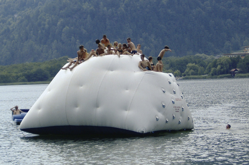 Eine Gruppe von Menschen sitzt und klettert auf einer großen aufblasbaren Struktur, die auf einem See schwimmt, mit Bergen und Bäumen im Hintergrund.