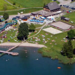 Luftaufnahme eines Erholungsgebiets am See mit Sandstrand, Steg, Wasserrutsche, Freibad, Sportplätzen, Rasenflächen und mehreren Gebäuden. Menschen nehmen ein Sonnenbad und schwimmen.