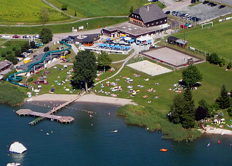 Luftaufnahme eines Erholungsgebiets am See mit Sandstrand, Steg, Wasserrutsche, Freibad, Sportplätzen, Rasenflächen und mehreren Gebäuden. Menschen nehmen ein Sonnenbad und schwimmen.