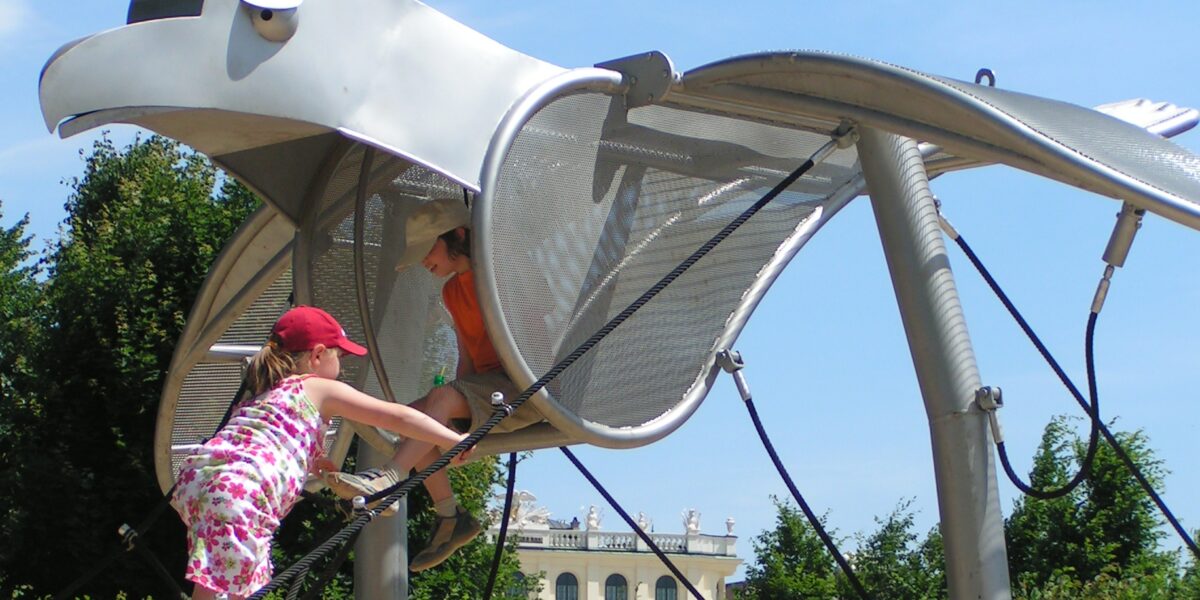 Zwei Kinder klettern und spielen auf einem großen Metallspielplatz in Form eines Vogels, im Hintergrund sind Grünflächen und ein Gebäude zu sehen.