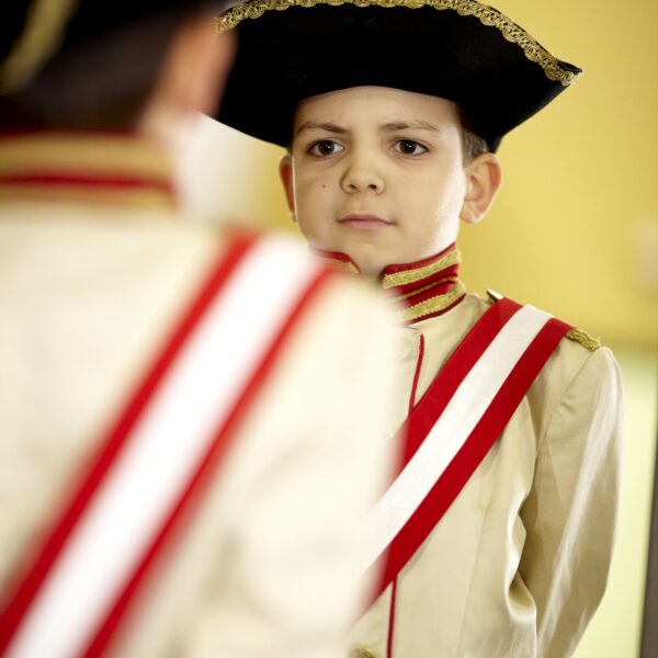Ein kleiner Junge in einer historischen Militäruniform mit Zweispitz und rot-weißer Schärpe steht vor einem Spiegel.