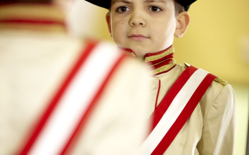 Ein kleiner Junge in einer historischen Militäruniform mit Zweispitz und rot-weißer Schärpe steht vor einem Spiegel.