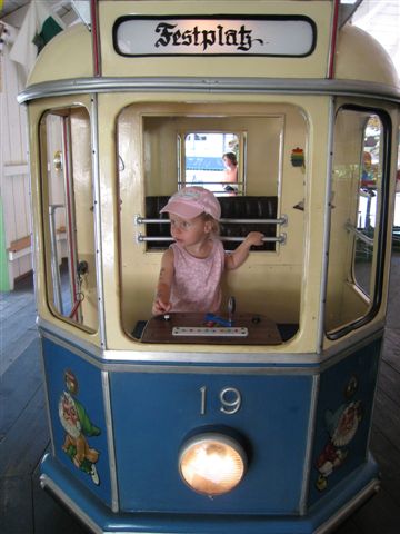 Ein kleines Kind mit einer rosa Mütze sitzt in einer blau-gelben Straßenbahn mit der Aufschrift "Festplatz" und der Nummer 19 und schaut zur Seite.