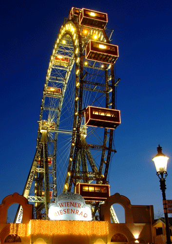 Das beleuchtete Wiener Riesenrad steht vor einem tiefblauen Abendhimmel, mit einem beleuchteten Eingangsschild und einer Straßenlampe im Vordergrund.