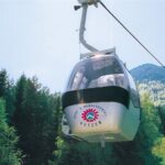 Eine weiße Seilbahn mit "Petzen"-Logo fährt über eine grüne, bewaldete Landschaft mit Bergen im Hintergrund.