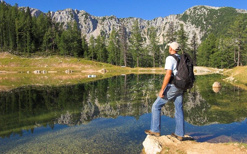 Ein Mann mit Rucksack steht auf einem Felsen an einem klaren See, umgeben von Bäumen und Bergen unter einem blauen Himmel.