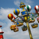 Buntes Fahrgeschäft im Vergnügungspark mit Sitzen in Form eines Heißluftballons, die sich in der Nähe eines rot-weiß gestreiften Turms vor einem teilweise bewölkten Himmel drehen.