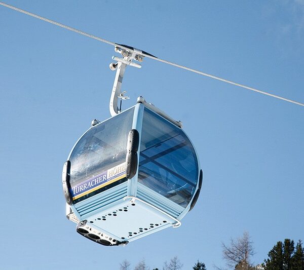 Eine mit blauem Glas umschlossene Gondelkabine hängt an einem Seil über Bäumen und einem teilweise bewölkten Himmel.