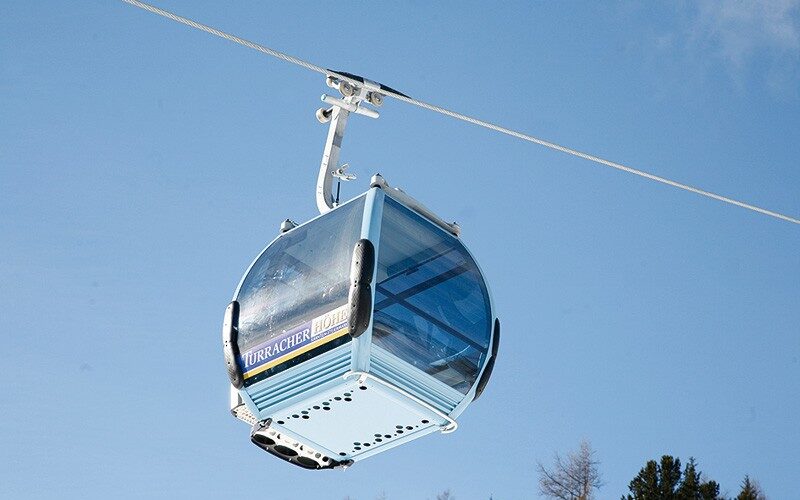 Eine mit blauem Glas umschlossene Gondelkabine hängt an einem Seil über Bäumen und einem teilweise bewölkten Himmel.