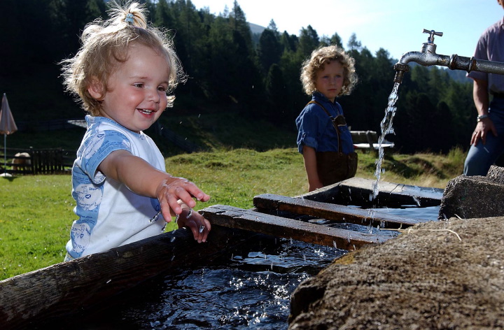 Ein kleines Kind lächelt, während es mit Wasser aus einem Wasserhahn im Freien spielt, während ein anderes Kind im Hintergrund auf einem grasbewachsenen Hügel steht.