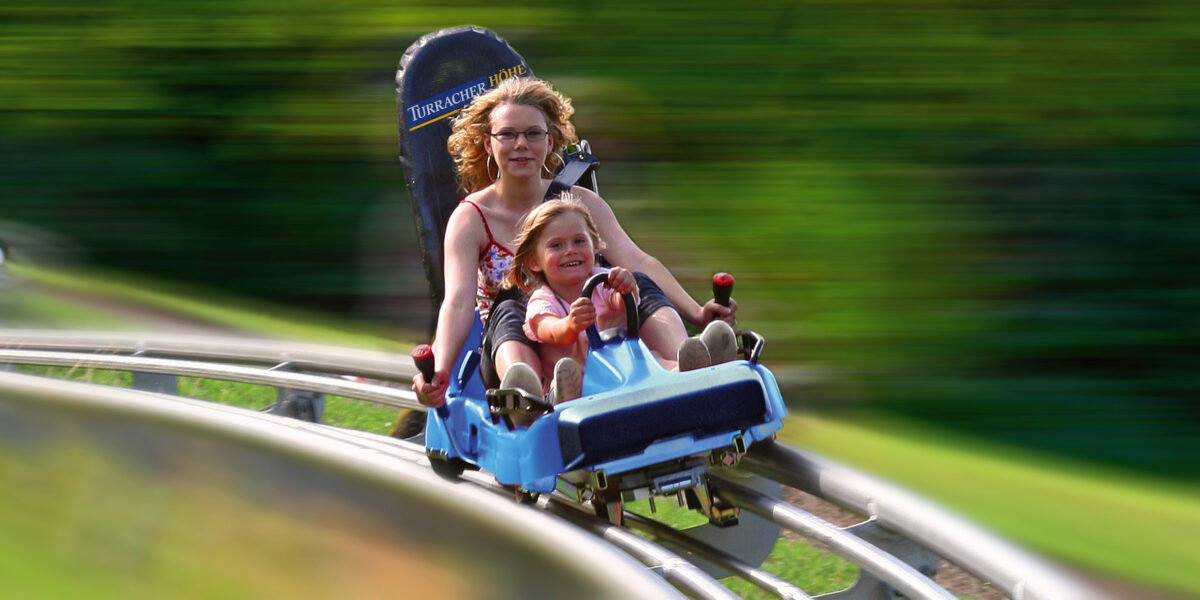Ein Erwachsener und ein Kind fahren gemeinsam auf einer blauen Alpenachterbahn, die über gebogene Gleise rasen, während das Grün im Hintergrund verschwimmt.