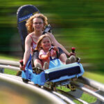 Ein Erwachsener und ein Kind fahren gemeinsam auf einer blauen Alpenachterbahn, die über gebogene Gleise rasen, während das Grün im Hintergrund verschwimmt.