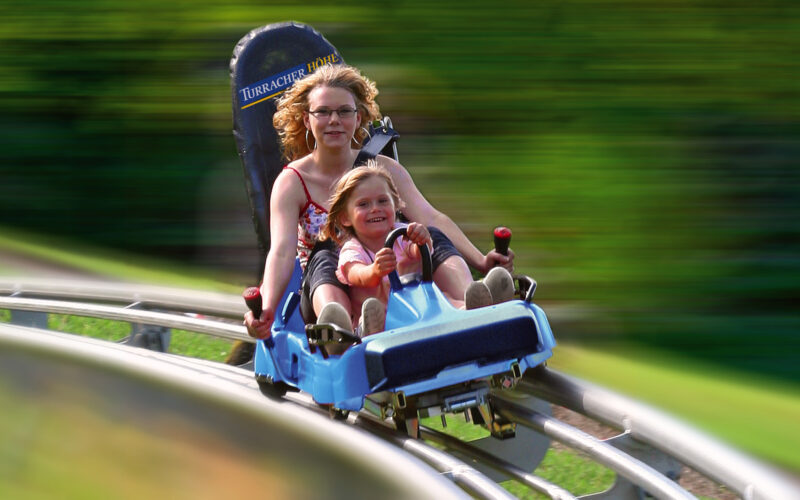 Ein Erwachsener und ein Kind fahren gemeinsam auf einer blauen Alpenachterbahn, die über gebogene Gleise rasen, während das Grün im Hintergrund verschwimmt.